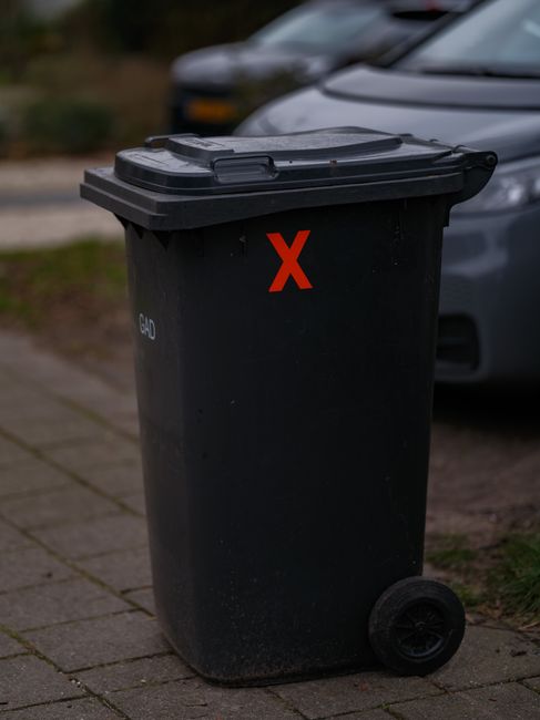 A black plastic wheelie bin with a closed lid, positioned on a paved sidewalk next to a parked car with a metallic grey body and dark windows. The bin features a red 'X' sticker on the front and appears to be used for waste collection, with a textured surface and sturdy design. The background shows a blurred street scene with other parked vehicles and some greenery, suggesting an urban or suburban residential area. The environment is outdoors during daytime with natural lighting, and the scene relates to private waste disposal options such as side or independent collection services. Waste Collection Holland Park likely offers rubbish removal solutions suited for home or commercial premises, as evidenced by the bin's placement in a typical driveway or street setting. The overall scene emphasizes urban waste management practices, including the use of personal waste bins for rubbish removal, in line with the company’s service category.