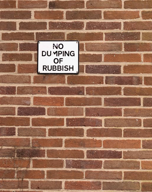 A rectangular metal sign with a white background and black lettering is mounted on a reddish-brown brick wall. The sign reads 'NO DUMPING OF RUBBISH' with the word 'DUMPING' partially obscured or faded, and the letters are in uppercase. The brick wall displays a typical running bond pattern with horizontal mortar joints, in tones of red, orange, and dark brown, with some variation in texture and coloration. The surface of the wall appears weathered and dry, with no visible vegetation or other objects nearby. The sign is positioned slightly above the center of the image, creating a clear focus for viewers, and the lighting is even, indicating daylight conditions. This scene suggests an area where waste removal or rubbish collection services might operate, with the sign implicitly discouraging illegal disposal of rubbish on-site or in the vicinity, aligning with principles of proper rubbish management by waste collection services such as Waste Collection Holland Park.