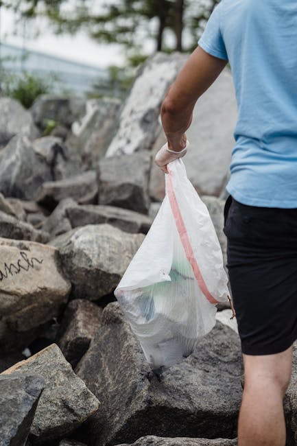 A person dressed in a light blue t-shirt and dark shorts is holding a white plastic rubbish bag with a red zip seal, collecting waste from a rocky outdoor area. The bag appears to contain various types of debris, possibly including plastic and paper materials, and is being lifted or moved away from the ground. The background features large, irregularly shaped rocks with some graffiti markings visible on one of the stones, and a blurred natural environment with trees and cloudy sky. The scene suggests an on-site waste clearance activity, emphasizing outdoor rubbish collection which could relate to private waste handling or alternative rubbish removal services offered by Waste Collection Holland Park, especially in contexts like environmental clean-ups or community collection efforts.