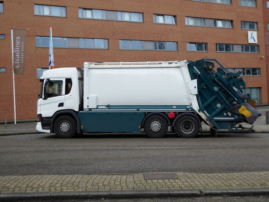 A large white and dark green waste collection truck parked on an urban street in front of a modern building with numerous rectangular windows. The truck is equipped with a hydraulic lift system at the rear, used for collecting and compacting waste materials, with visible mechanical arms and containment mechanisms. The vehicle's cab is positioned at the front, featuring side mirrors and a window, while the body of the truck extends along the length of the street, indicating its purpose for waste and rubbish removal services. The surrounding environment includes a paved road with a brick-patterned sidewalk, and a flagpole with a vertical sign indicating a hotel or residential complex in the background. The scene is lit with natural daylight, and the overall setting appears clean and organized, reflecting a typical waste collection operation that might be associated with private rubbish removal services, such as those offered by Waste Collection Holland Park.