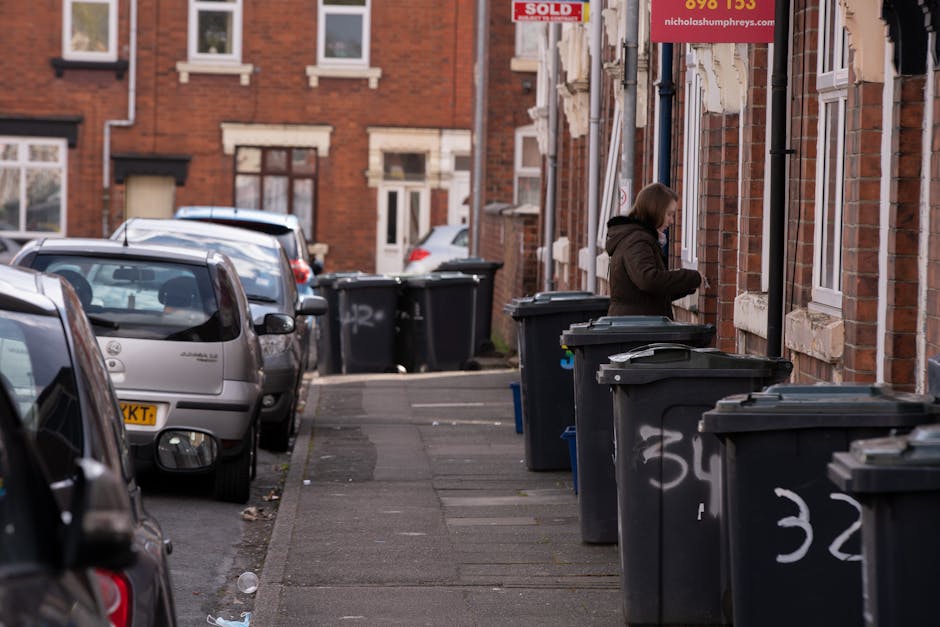 A narrow residential street lined with parked cars on the left, including a silver hatchback and a dark-colored vehicle, with a sidewalk on the right side. Multiple black wheelie bins are positioned along the sidewalk, some with white painted numbers on their sides. A woman wearing a brown coat is seen standing next to the bins, opening or examining one of them. The background features brick terraced houses with white framed windows and doors, some with small front garden areas or steps. There are signs attached to the buildings, one red advertising a property sale and another partially visible in the upper right corner. The scene is lit with natural daylight, casting soft shadows and highlighting the clean, orderly arrangement of waste containers in an urban environment, which aligns with private waste collection or rubbish removal services like Waste Collection Holland Park.
