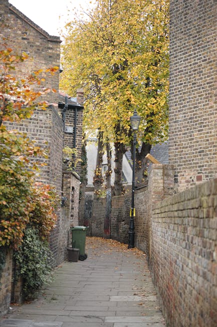 A narrow paved alleyway in an urban setting, bordered by tall brick walls on both sides. The left wall features some climbing foliage with orange and green leaves, while the right wall is composed of weathered brown bricks. A green wheeled waste bin is positioned near the corner on the left side, close to the base of a large deciduous tree with yellow and orange autumn leaves, which extends over the alleyway. A traditional black lamppost with a glass lantern is situated along the right side, slightly behind the bin, and a small wooden plank is leaning against the wall further down the alley. The environment suggests an older part of a city or town, typical of areas where independent waste collection or on-site clearance services like those offered by Waste Collection Holland Park might be employed for rubbish removal or disposal. The scene is illuminated by natural daylight, highlighting the seasonal foliage and textures of the brick walls and paving stones, creating a quiet, residential atmosphere associated with private waste handling or alternative rubbish collection methods.