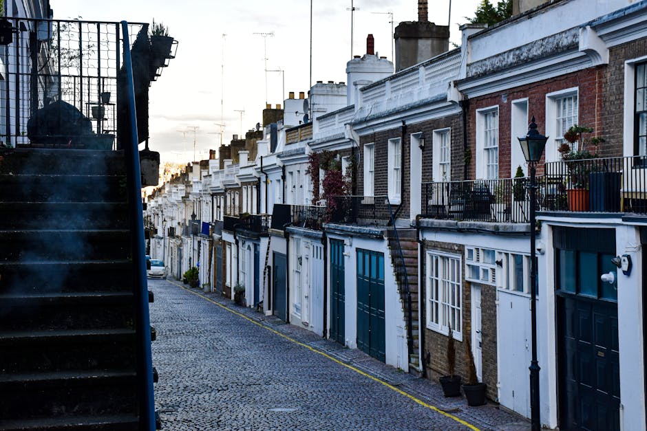 The image depicts a narrow cobblestone street lined with a series of terraced houses that feature traditional brick facades, some partially painted white and others showcasing exposed red brickwork. Each residence has a small front balcony enclosed with black metal railings, some adorned with potted plants and outdoor decorative items. The houses have white window frames and various front doors painted in shades of dark green and blue, with some doors equipped with modern doorbells and outdoor lighting fixtures. On the left side of the image, a dark metal staircase with a grated railing ascends to an elevated entrance, partially obscuring the lower part of the street. The background shows a sky transitioning from daylight to dusk, with faint silhouettes of television antennas and chimneys on the rooftops, suggesting an urban residential setting. The scene emphasizes a quiet, well-maintained street typical of inner-city environments where private waste collection services, such as those from Waste Collection Holland Park, may be employed for rubbish removal and on-site clearance, avoiding reliance on municipal waste systems.
