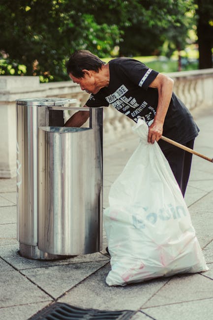 A middle-aged man with dark, combed-back hair is seen disposing of rubbish into a modern, stainless steel public waste bin on a paved sidewalk. He is wearing a black short-sleeved T-shirt with white graphic or text elements on the front and dark trousers. Standing next to the bin, he holds a large white plastic bag filled with waste using his left hand, while his right hand is reaching into the opening of the bin, which has a rounded, polished finish. The background features lush green foliage and trees, suggesting a park or urban outdoor area. The scene is lit with natural daylight, highlighting the reflective surface of the bin and the textures of the pavement, which is composed of square concrete slabs. The image subtly relates to private waste handling and alternative rubbish collection methods, exemplifying responsible disposal practices.