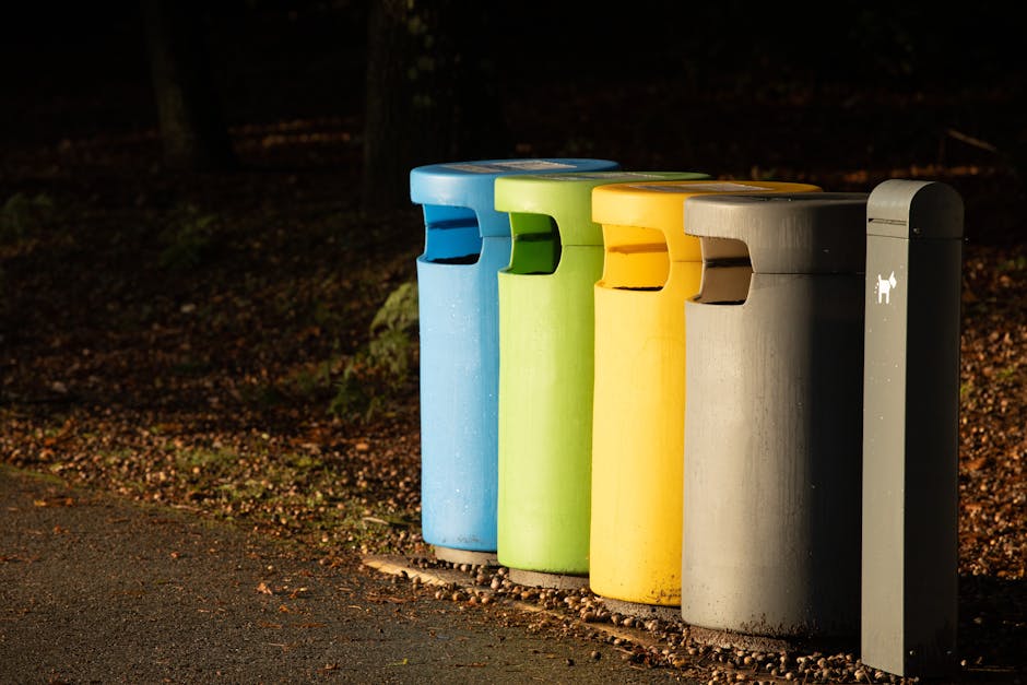A collection of overflowing rubbish bins and discarded waste bags is situated on a paved sidewalk in front of a commercial building, with a metal railing partially enclosing the area. The waste includes black, red, and green bins, some with lids open, revealing mixed household and packaging waste like cardboard boxes, plastic bags, paper, and discarded packaging materials. Some cardboard boxes and paper products are scattered on the ground around the bins, with a variety of textures from smooth plastics to corrugated cardboard and crinkled paper. Behind the waste, a building with contemporary glass and metal façade features large windows and colorful signage, likely indicating shops or eateries, with a scaffolding structure visible on the upper level. A parked silver car with a yellow license plate is adjacent to the waste area, and two small trees with bare branches frame the scene on the left. The environment appears to be an urban commercial or retail area, and the current state of the waste suggests an on-site collection or an overflow from nearby refuse management services. Waste Collection Holland Park offers services relevant to this scene, highlighting the importance of proper rubbish disposal and alternative waste handling options. The overall scene is well-lit by natural daylight, emphasizing the cluttered and unmanaged appearance of the littered waste.
